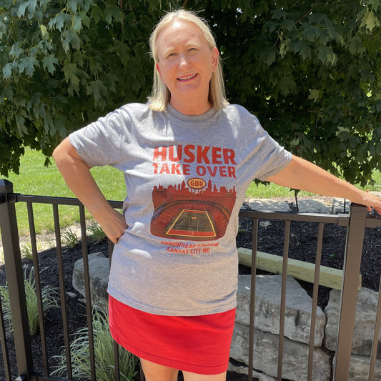 Person wearing a gray 'Husker Take Over' t-shirt and red skirt on a wooden deck with greenery in the background.